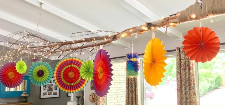 DIY branch chandelier wrapped with string lights and colorful paper decorations hanging indoors