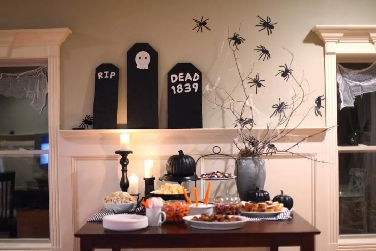 Halloween buffet table with tombstone backdrop, spider wall decor, and black pumpkins