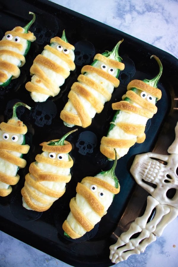 Jalapeño poppers wrapped in dough like mummies with candy eyes on a black tray