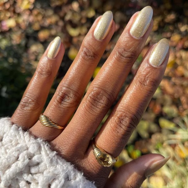Hand with short gold shimmer nails shown outdoors, wearing cozy cream sweater and gold rings.