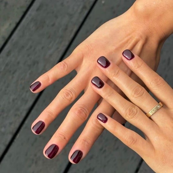Short burgundy nails shown on both hands with a glossy finish and simple gold ring.