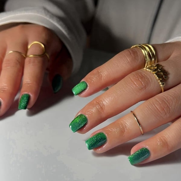 Square green shimmer nails with a sparkly finish, styled with layered gold rings.