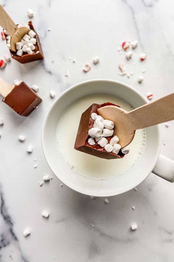 block of chocolate covered in mini dried mashmallows attached to the end of a wooden spoon about to be dipped into hot milk to make hot cocoa