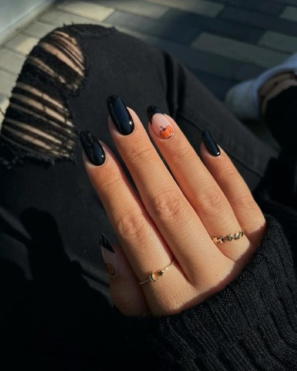 Glossy black almond-shaped nails with a nude accent nail featuring a small orange pumpkin.