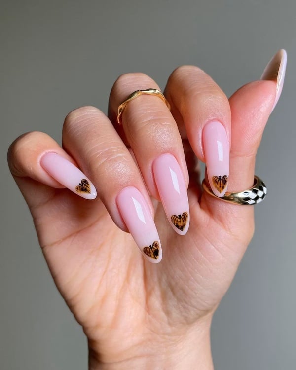 Pale pink almond-shaped nails with small brown and amber heart-shaped pumpkin designs at the tips.