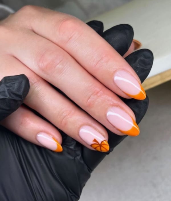 Nude almond-shaped nails with bright orange French tips and a small orange pumpkin accent along one tip.