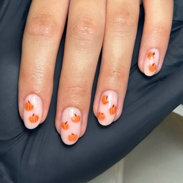Short pale pink nails decorated with multiple tiny orange pumpkins across each nail.